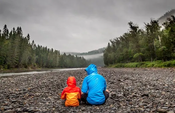 Parent et enfant près d'une rivière entourée de forêts de sapins